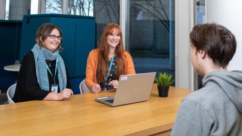 Kuvassa kolme henkilöä. Kaksi istuu pöydän toisella puolella kasvot kuvaajaan päin ja yksi selin. Kaikki hymyilevät. Heidän välissään on tietokone ja menossa on jonkinlainen ohjaustilanne.