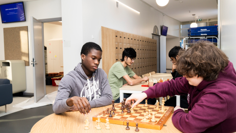 Four people are playing chess, two in the front and two in the back, using the chessboard on the table.