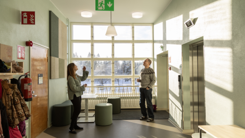 Kaksi henkilöä katsoo ylöspäin kohti kattovalaisimia koulun käytävän päässä ikkunan edessä.