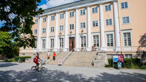 Wide steps lead up to a large light yellow building. People are walking in front of the building. The weather is sunny.
