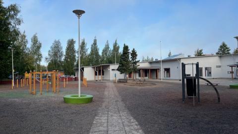 Metsäniityn päiväkodin ja koulun pihaa, taustalla näkyy rakennus. Piha on lähinnä avointa sorakenttää, jossa on puisia leikkivälineitä. on 