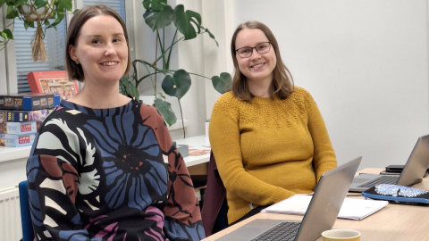 Kuvassa kaksi naista istumassa pöydän ääressä. Molemmat hymyilevät.