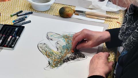 Harmaahiuksinen nainen maalaa värikkäillä liiduilla abstraktia teosta paperille.