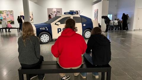 Kolme henkilöä istuu penkillä museossa. Taustalla teoksia ja poliisiautoteos.