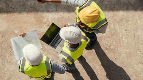 Kolme rakennusalalla työskentelevää henkilöä seisoo pihalla. Heidät on kuvattu ylhäältä päin. Päässä kypärät ja päällä huomioliivit. Yksi osoittaa kädellään eteenpäin, toisella on sylissä läppäri ja kolmannella papereita.