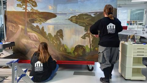 Kaksi mustapuseroista henkilöä tarkastelee isoa maisemamaalausta. 