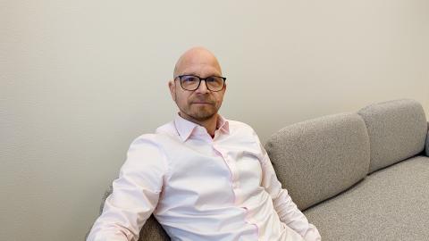 Risto is sitting on a light grey sofa wearing a pink collared shirt and looking straight at the camera.