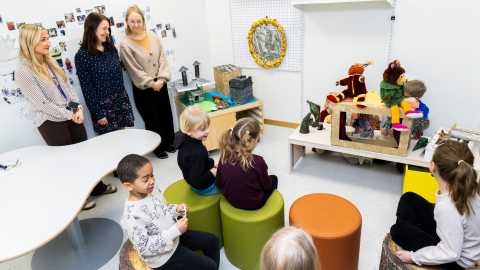 Lapset esiintyvät edessä ja toiset katsovat istuen. Päiväkodin työntekijät seuraavat tilannetta seisoen.