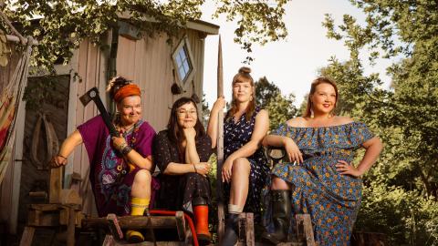 Four smiling women pose for a photographer. They are wearing rubber boots, one holding an axe and the other a fence post.
