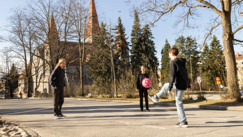 Kolme nuorta potkii asfaltilla palloa. Takana on kirkko.