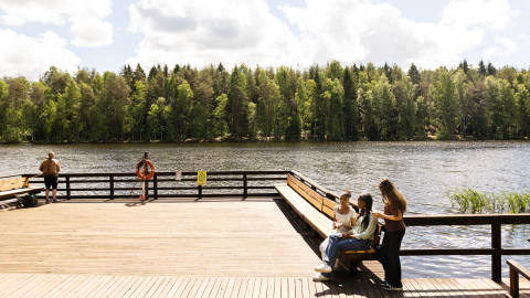 Yksi mies kalastamassa ja kolme nuorta tyttöä istumassa Ahvenisjärven uudistetulla laiturilla poutaisena kesäpäivänä. 