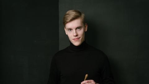 Kapellimestari Kristian Sallinen mustassa poolossa, tahtipuikko kädessään mustaa taustaa vasten.