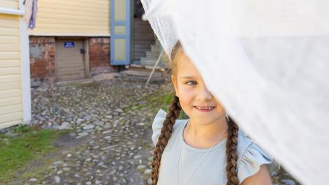 Pikkutyttö jurkistaa valkoisen lakanan takaa. Taustalla Amurin museokorttelin taloja.