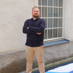 Mies seisoo kädet puuskassa vaalean rakennuksen seinämällä, taustalla ruutuikkuna.