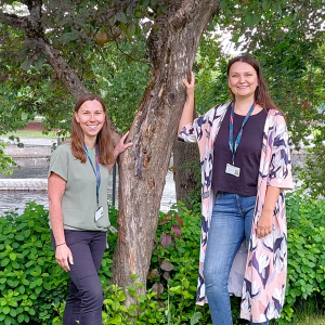 Kaksi hymyilevää naista nojaa puuhun, yksi kummallakin puolella.