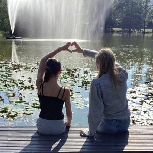 Kaksi tyttöä istuu Sorsapuiston lammen rannalla ja muodostaa sormilla yhdessä sydämen. 