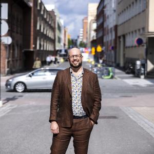 Mies seisoo kadulla hymyillen ja katsoo kameraan. 