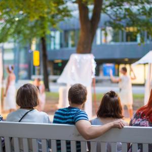 Viisi henkilöä istuu Hämeenpuistossa penkillä ja katsoo edessä olevaa taide-esitystä.