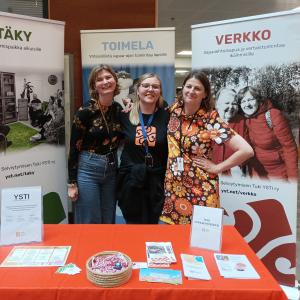 Kolme naista seisoo hymyillen esittelytiskin takana messutilaisuudessa.