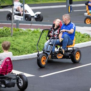 In a traffic park, children pedal on a pedal cars, in the middle  a parallel pedal car with a child and an adult.