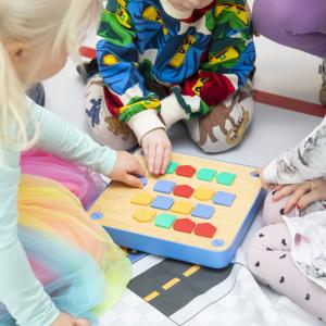 Kolme lasta istuu lattialla ja pelaa lautapeliä, jossa on värikkäitä nappuloita.