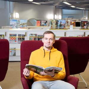 Henkilö istuu nojatuolissa pääkirjasto Metson lehtisalissa ja pitää käsissään avointa lehteä. Taustalla on salin lehtikalusteita. 