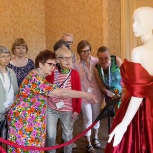 Naiset seisovat museonäyttelyssä ja katselevat punaista, olkaimetonta silkkisatiinista valmistettua iltapukua.