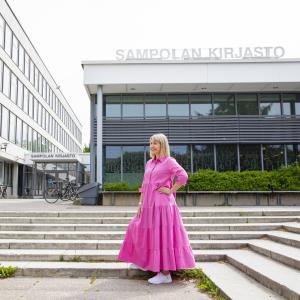 Henkilö seisoo vaaleanpunaisessa pitkässä mekossa rakennuksen edessä, jossa lukee Sampolan kirjasto.