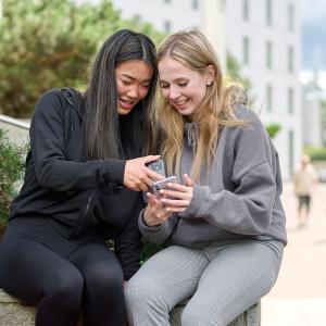 Kaksi nuorta naista istuu Tampere-talon lähellä katsomassa iloisesti puhelinta.