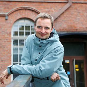 Mikko katsoo suoraan kameraan taustallaan Frenckellin punatiilinen rakennus.