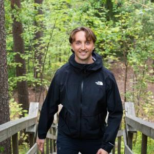 Henkilö seisoo hymyillen puuportaiden yläpäässä vehreän metsämaiseman keskellä.