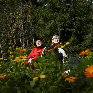 Kaksi henkilöä istuu keskellä yhteisöviljelmää, ympärillä kukkia, taustalla metsää.