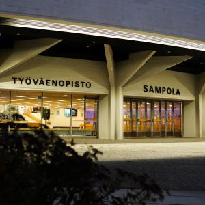 Sampola-rakennus pimeällä. Seinä on valaistu. Etualalla on pensas.