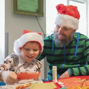 Pieni tyttö ja mies askartelevat korttia iloisina tonttulakit päässään.