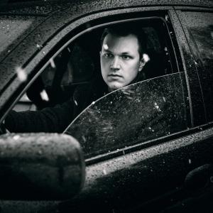 Mustavalkoisessa kuvassa Harry-Pekka on sateisen auton sisällä, etuoven lasi auki.