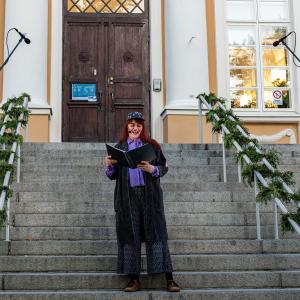 Nainen seisoo portailla ja lukee käsissään pitelemästään kansiosta runoa.