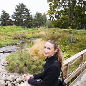 Tummatakkinen henkilö seisoo sillalla ihaillen ojaa, joka sijaitsee kauniissa ympäristössä.