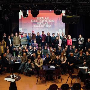 Suuri ryhmä ihmisiä poseeraa yhteiskuvassa. Taustalla valotaulu, jossa lukee &quot;Nekalan kulttuurikeskus Nekalab avaa ovensa 1.1.2026&quot;.