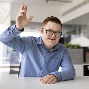 Kauluspaitaan pukeutunut henkilö, jolla on Downin oireyhtymä, nostaa toista kättään.