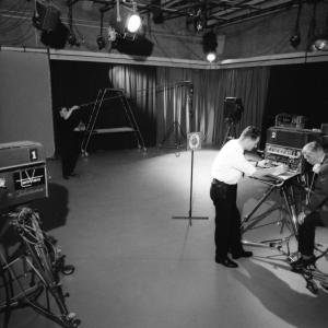 Kaksi henkilöä seisoo televisiolaitteen ääressä studiossa. Etualalla on videokamera.