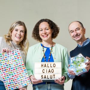 Kolme henkilöä seisoo vierekkäin. Heillä on käsissään kielten opiskelutavaroita, kuten lippuja ja taulu.