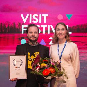 Palkinnon voittaja poseeraa kukkien ja taulun kanssa kaupungin edustajan kanssa. 