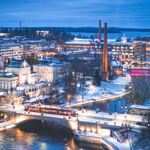Ilmakuva Tampereen keskustasta talvella sinisessä hämärässä. Kaupungin valot loistavat kirkkaasti. Hämeensillan yli kulkee ratikka.