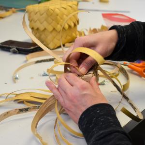 Henkilö sitoo tuohisuikaleita pöydän ääressä. Taustalla tuohirasia.