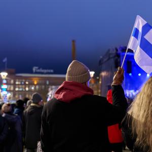 People in the evening at the winter time in the Central Square, with a person holding a Finnish flag. Finlayson building in the background.