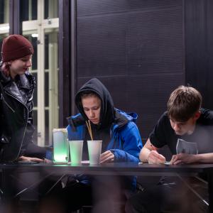 Kaksi nuorta istuu pöydän ääressä kirjoittamassa ja yksi nuori istuu pöydällä ja hymyilee