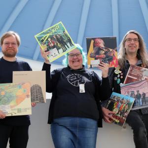 Kolme henkilöä seisoo iloisena vinylilevyjä käsissään.