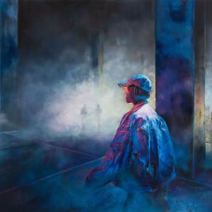 Nuori lippispäinen mies seisoo hämyisessä, tyhjässä tehdashallissa. 