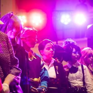 Lavalla värivalojen loisteessa cosplay asuihin pukeutuneita ihmisiä rivissä muotinäytöksessä..