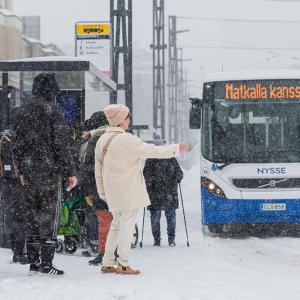 Talvisella pysäkillä nainen pysäyttää bussin, jossa lukee: Matkalla kanssasi.